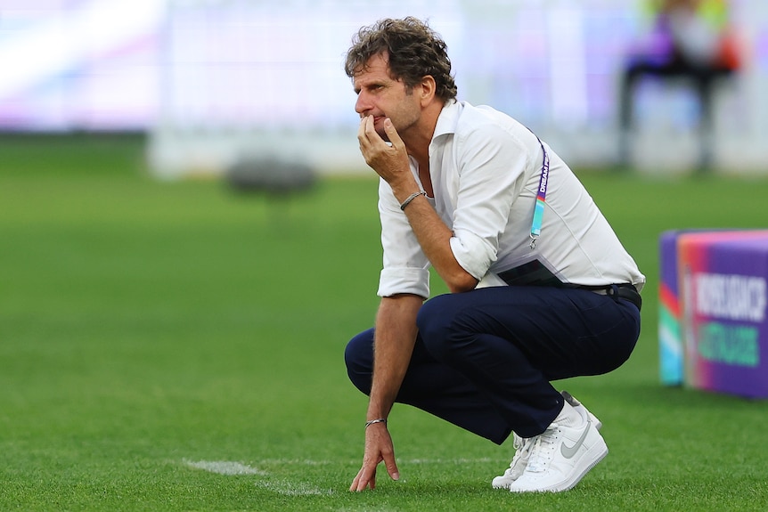Matildas coach Joe Montemurro is squatting down with hand on chin, watching on the sidelines.