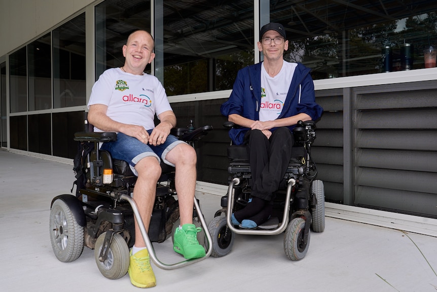 Berrie in a white top and shorts in a wheelchair, Marcel in a cap and hoodie in a wheelchair outdoors