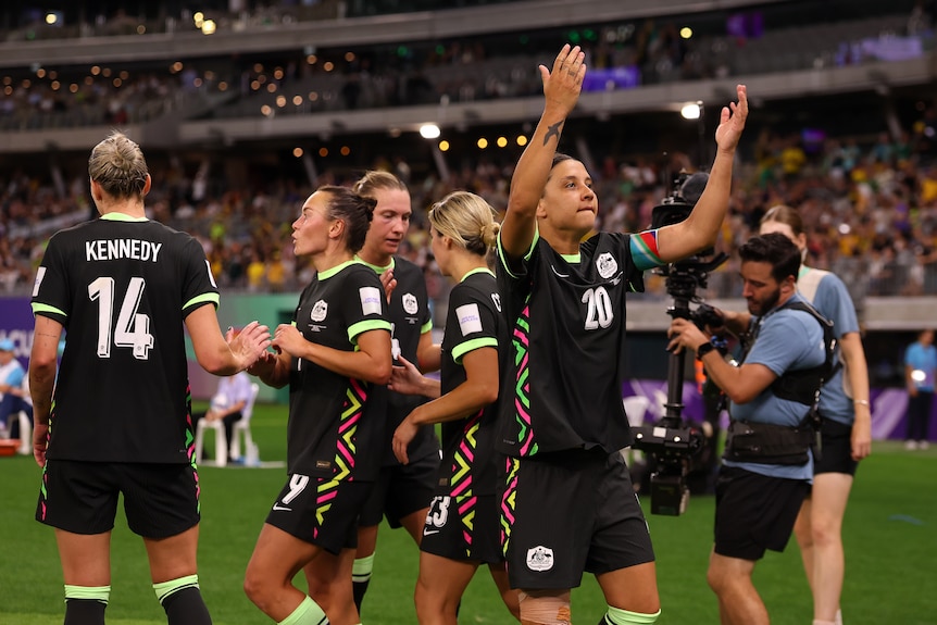 A soccer player in green holds up her hands in the air with teammates around her