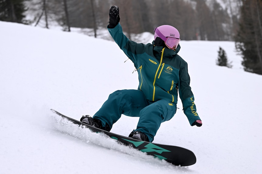 A snowboarder wearing the green and gold uniform for Australia and a pink helmet is leaning into a turn on the slopes.