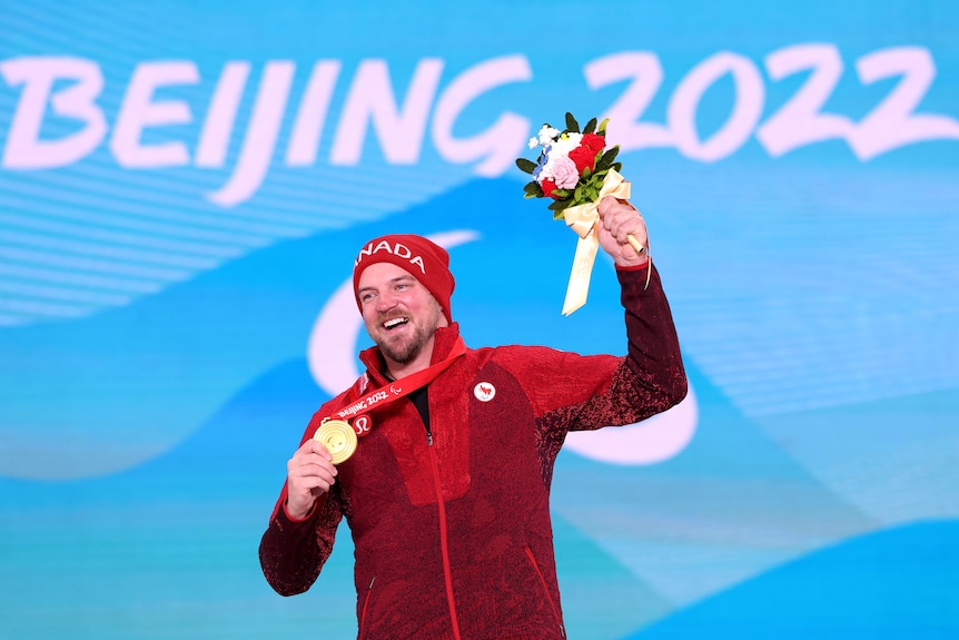 A man wearing the Canadian uniform and beanie, is grinning as he holds up his gold medal and bouquet.