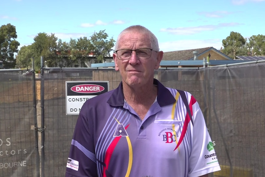 Man wearing purple, white, red and yellow polo shirt, glasses, standing in front of a construction site gate.