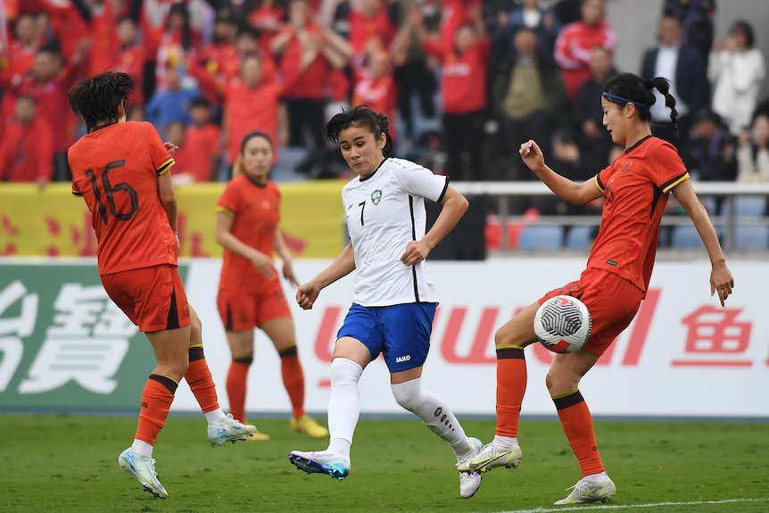 A member of the Uzbekistan women's football team has passed the ball as two defenders approach to tackle.