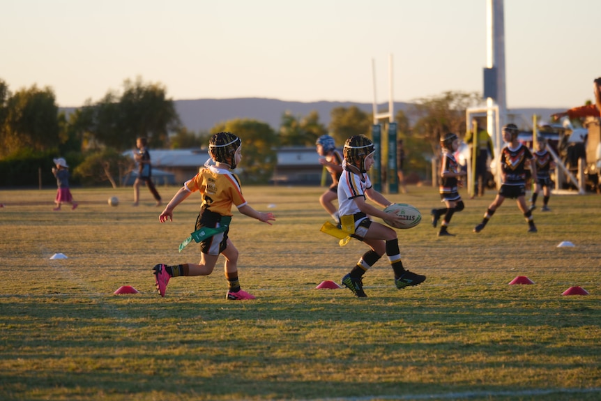 Two young children in rugby uniforms run across a grass pitch at sunset.