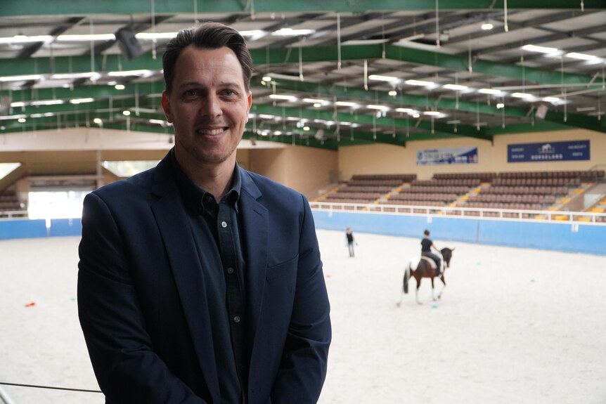 A man smiles to camera in an equestrian facility.
