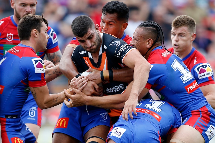 A Wests Tigers NRL player is tackled by several Newcastle Knights opponents.
