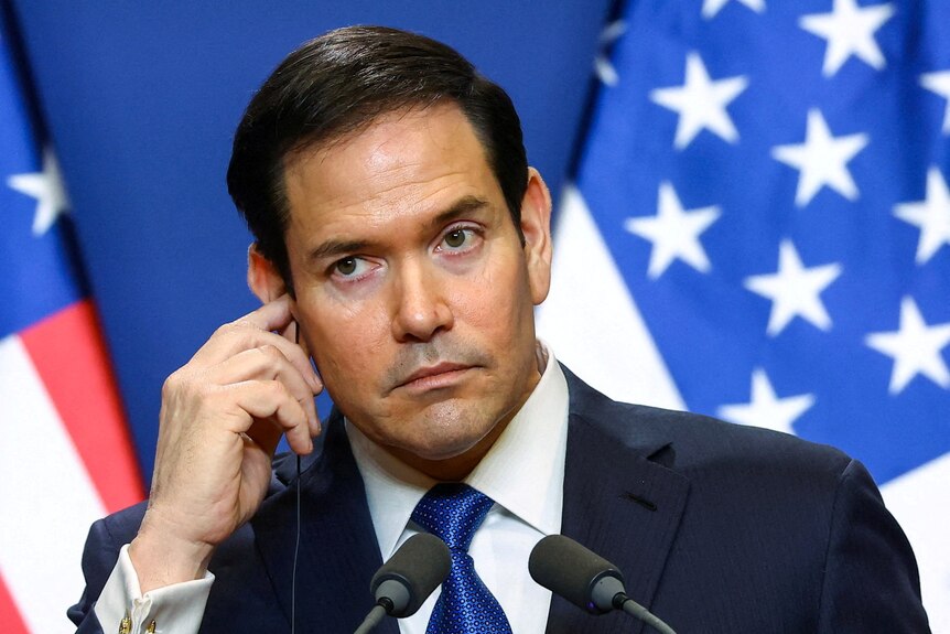 Marco Rubio touching his right ear while standing emotionless in front of a US flag.