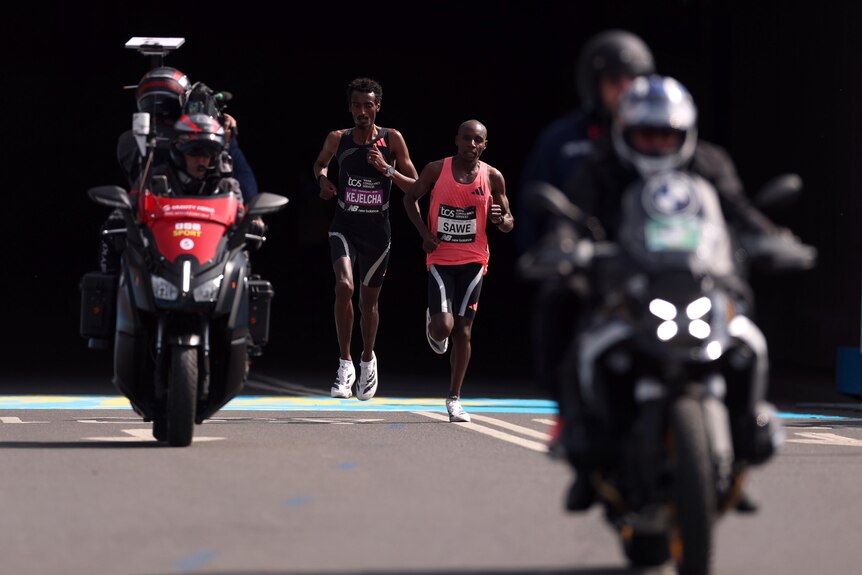 Sabastian Sawe runs with Yomif Kejelcha in a marathon event, flanked by motorcycles
