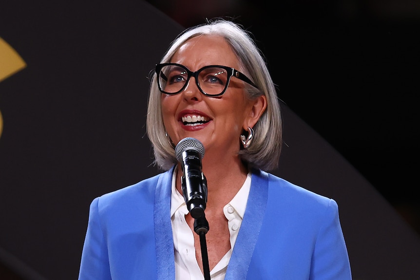 Liz Ellis speaks in front of a microphone in a blue suit