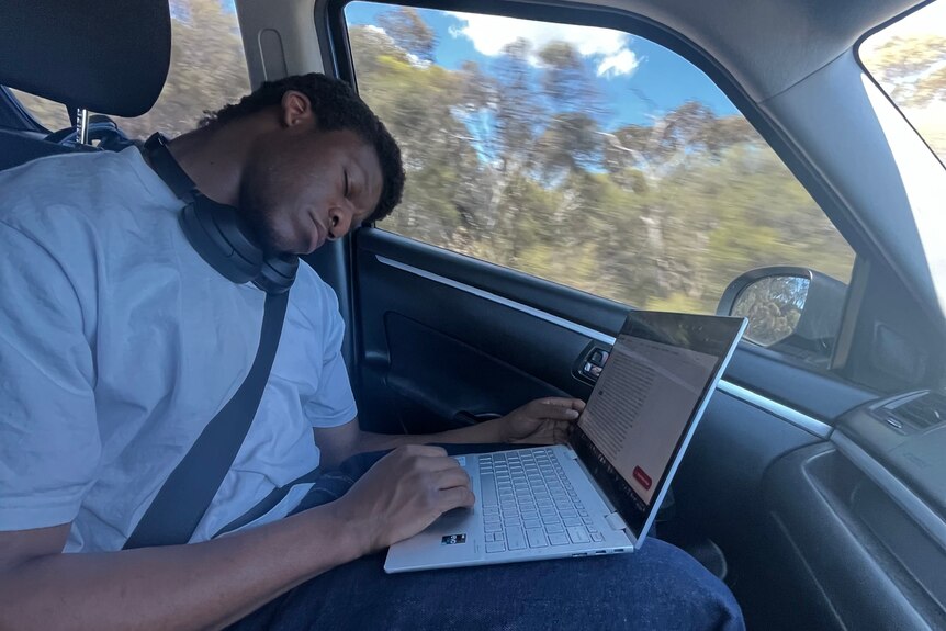 A man in a t-shirt sleeping in the passenger seat of a car with his laptop in his lap.