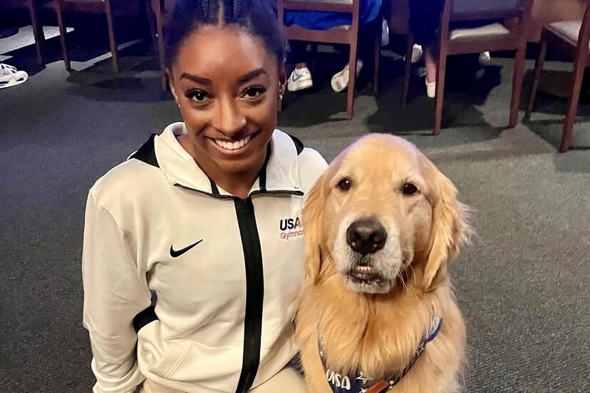 Simone Biles crouches down and smiles next to a golden retriever
