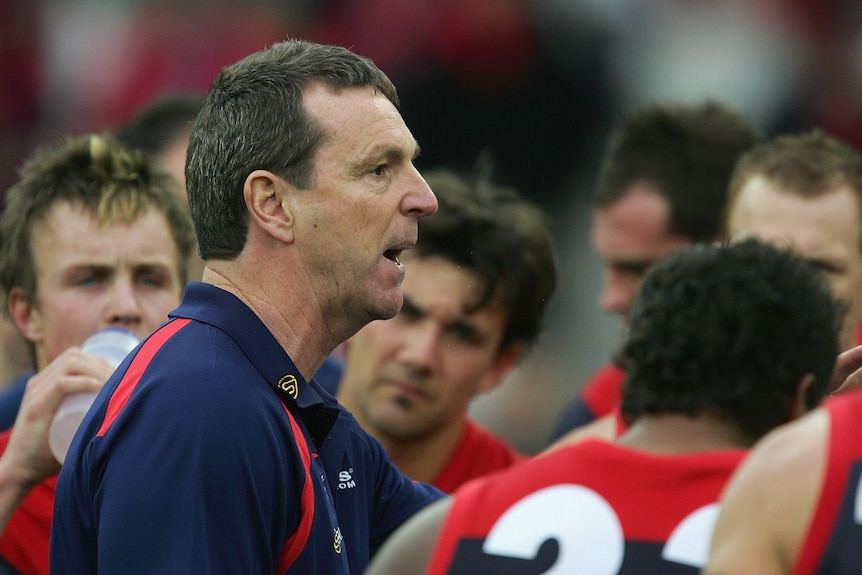 A man in a polo short talks to a huddle of football players. 