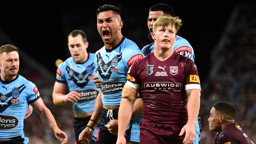 A NSW State of Origin player roars in celebration as a Queensland player looks dejected after a try.