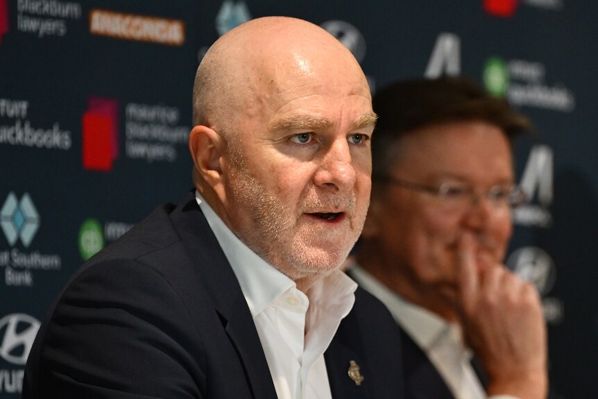 Graham Wright speaks at a press conference, is bald, wears a black suit, white shirt.