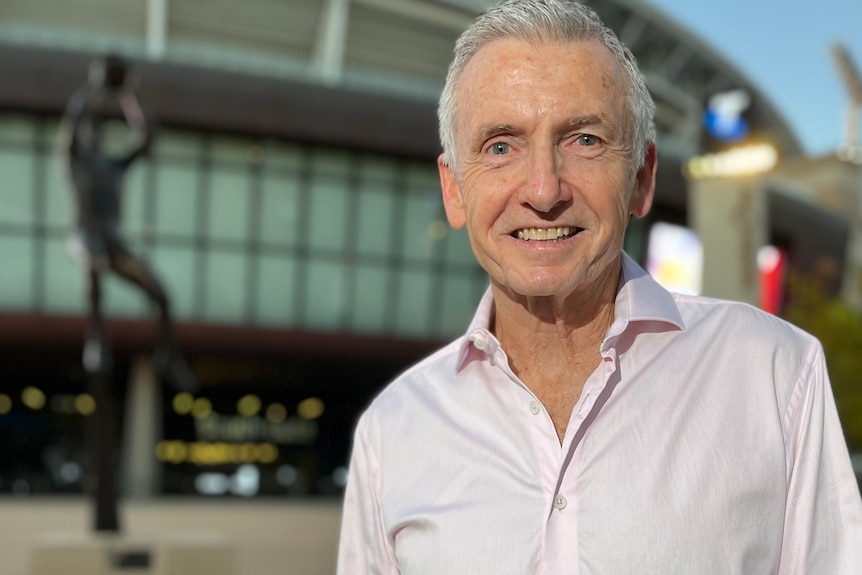 A man in a white shirt and with grey hair stands outside a football stadium.