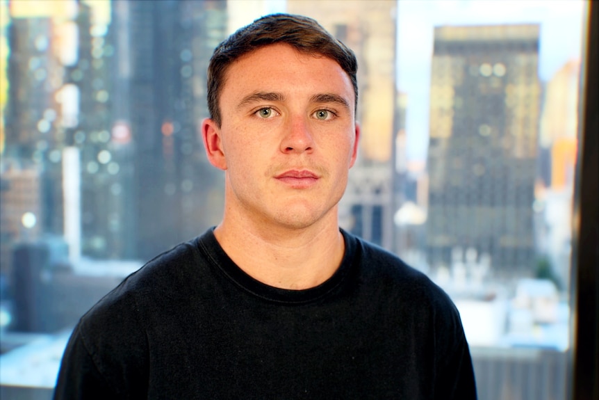 Aiden O'Driscoll stands in an office with the city skyline behind him, looking serious.