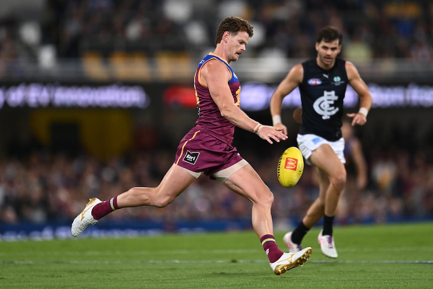 A Brisbane Lions AFL player draws his foot back to kick the ball downfield, as a Carlton player looks on.