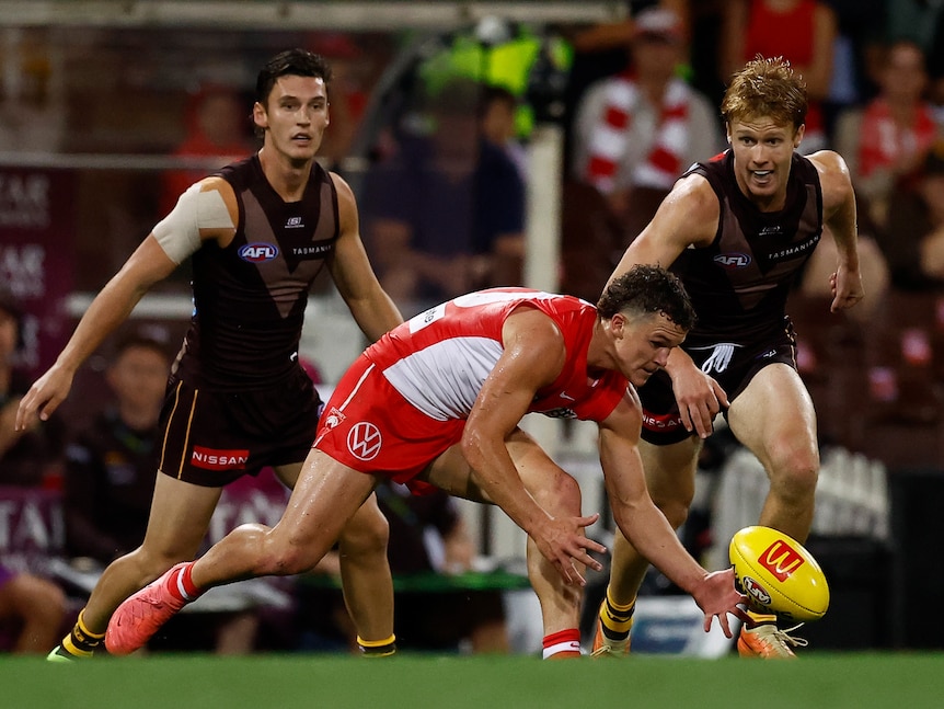 A Sydney AFL player bends down to pick up the ball as two Hawthorn players close in.