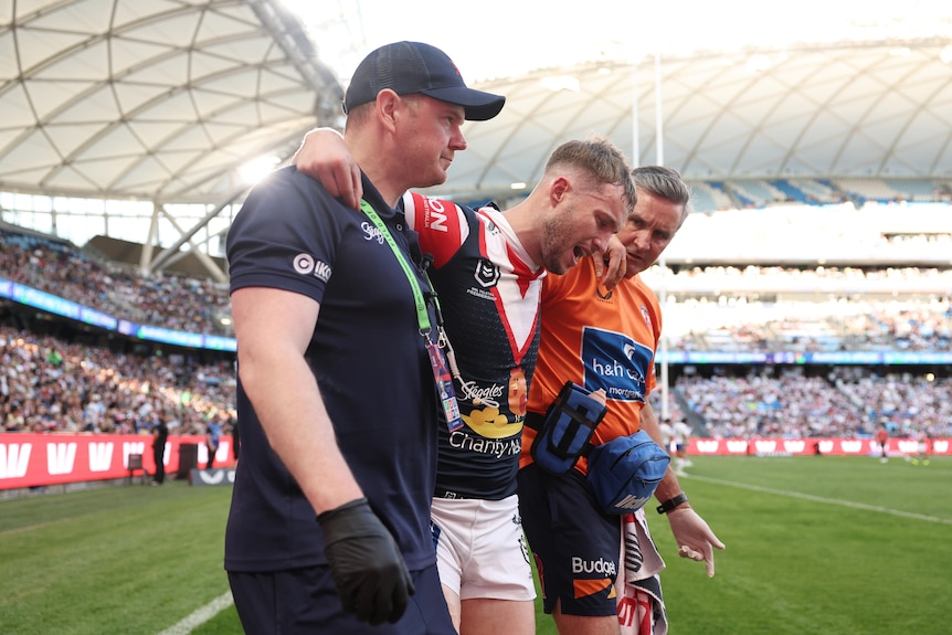 Sam is taken from the field during an NRL match, arm in arm with two men, one in all black.
