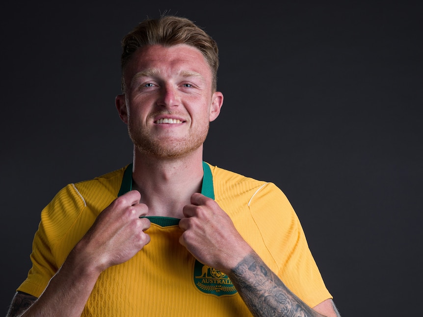 Harry is in yellow and green jersey, holds collar, smiles, has tattoos on his left arm. Background dark.