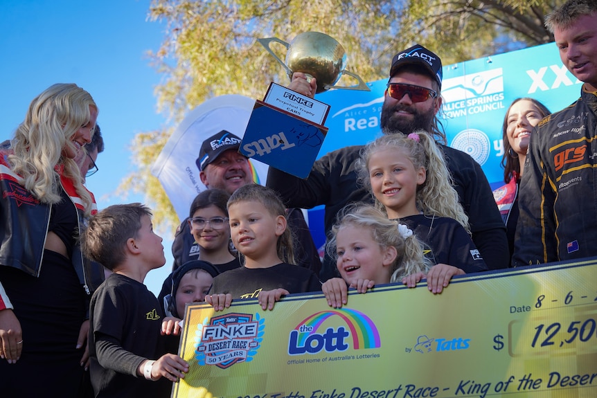 A group of people holding up a large competition winner's cheque, as one man in the group holds up a large gold trophy.