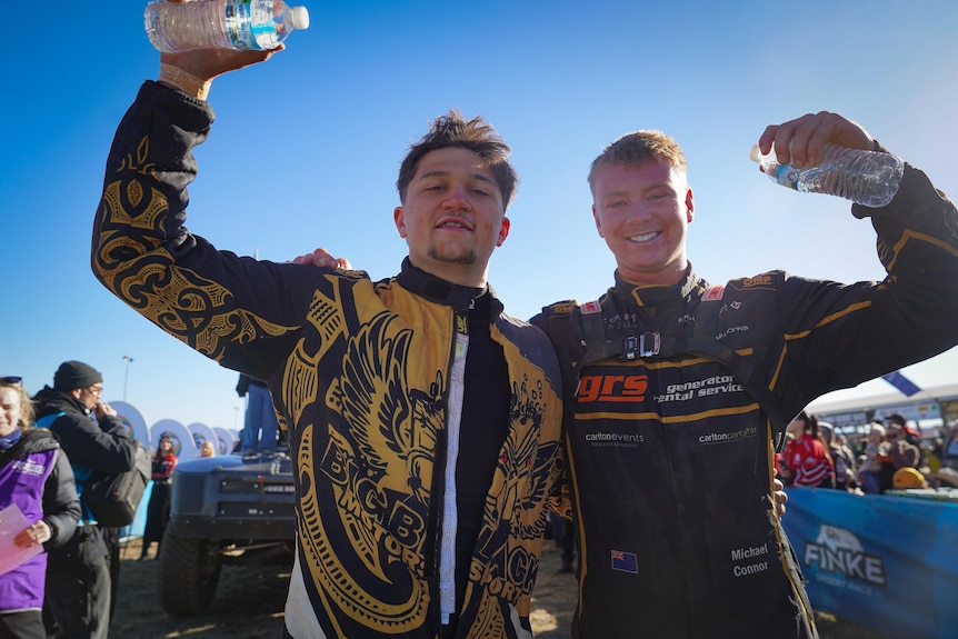 Two young men in car racing jackets standing next to a race track, smiling and with their arms raised triumphantly.