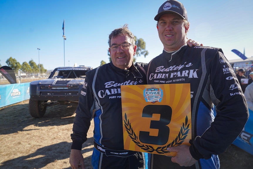 Two men standing next to each other and smiling, holding a large piece of cardboard with "3" on it, beside a car racing track.