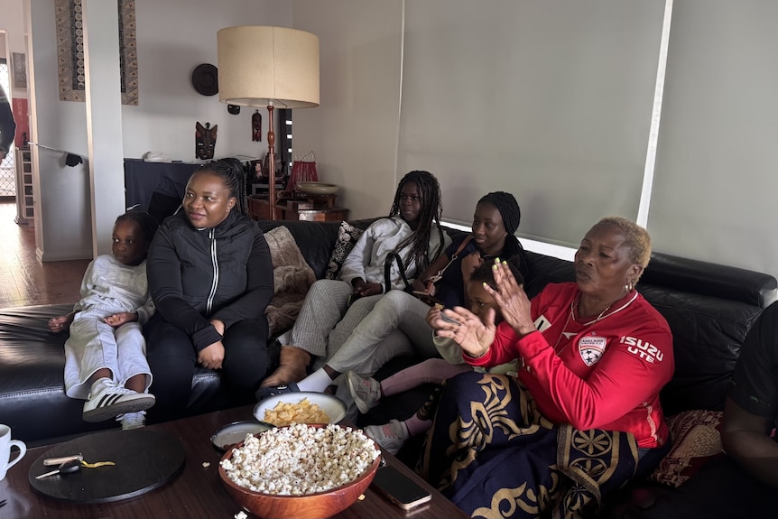 A family gather in a living room clapping as they watch TV