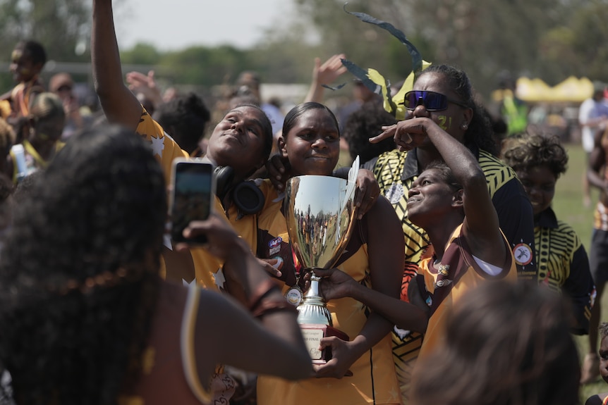 A group of women in yellow jerseys, with one in the centre holding a large gold trophy