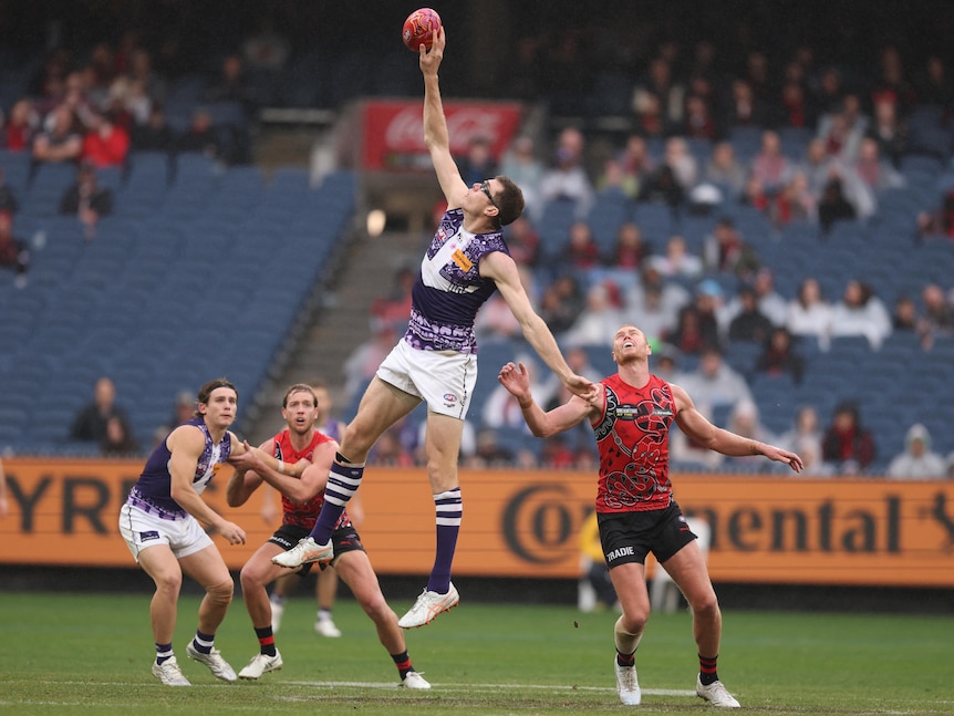 A Fremantle AFL ruckman leaps high to tap the ball as an Essendon opponent stands watching.