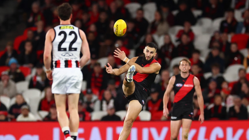An Essendon player extends his leg as he kicks through the ball to send it downfield as a St Kilda defenders stands on tiptoe.