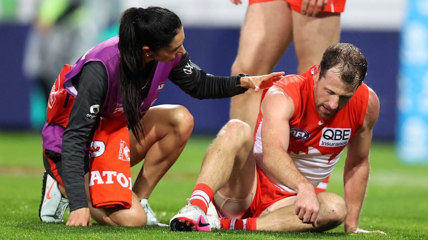 A Sydney AFL player sits dazed on the ground as a trainer puts her hand on his shoulder to check on him.