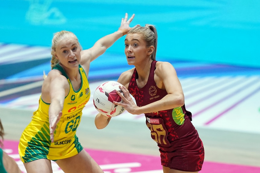 A female netballer holds the ball and looks to pass as another netballer from a different team holds her arms out defending.