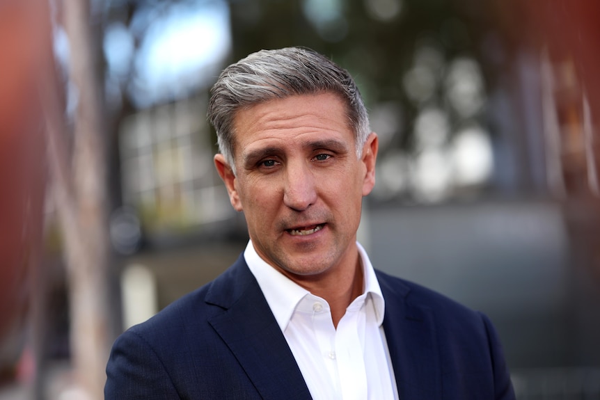 Man in a white shirt and navy suit jacket at a press conference.