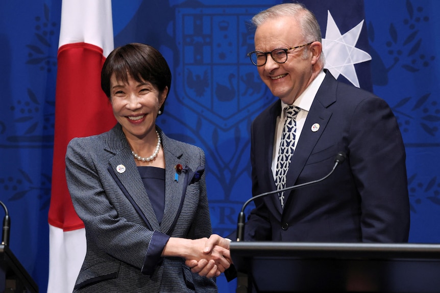 A woman in a blazer and a man in a suit shake hands and smile widely in front of the Japanese and Australian flags.