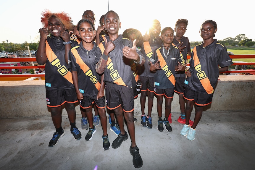 Smiling boys and girls in dark sports outfits give the thumbs up while standing on a concrete walkway in a stadium.