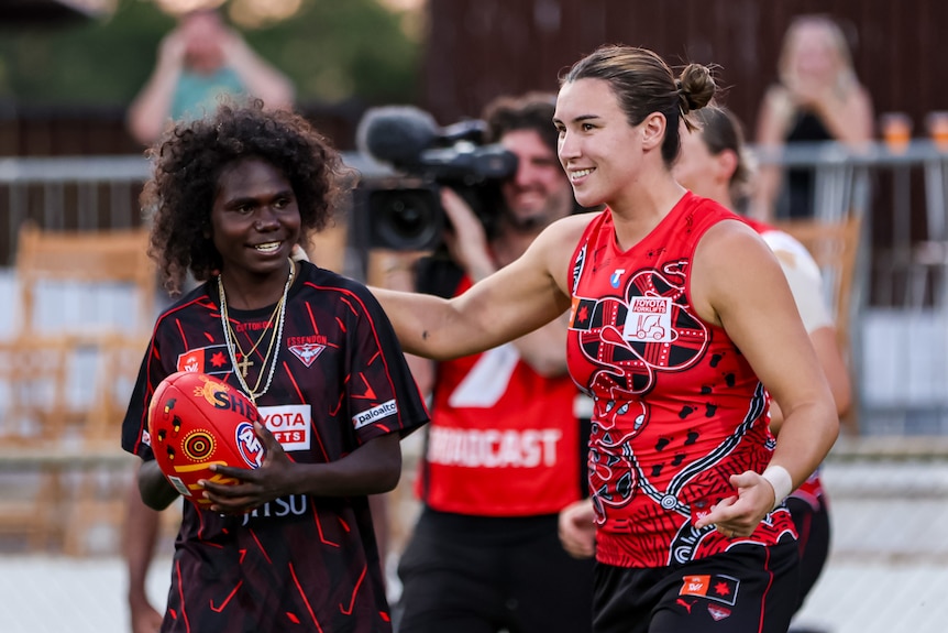 Two smiling women, one in an AFLW uniform, stand together on an oval.