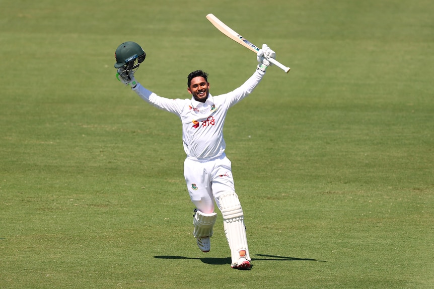 A cricket batter holds his helmet and bat to the sky, smiling and celebrating a century