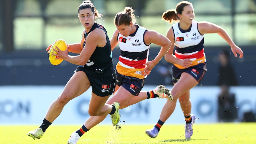 A Carlton AFLW player runs with the ball while being chased by two Adelaide defenders.