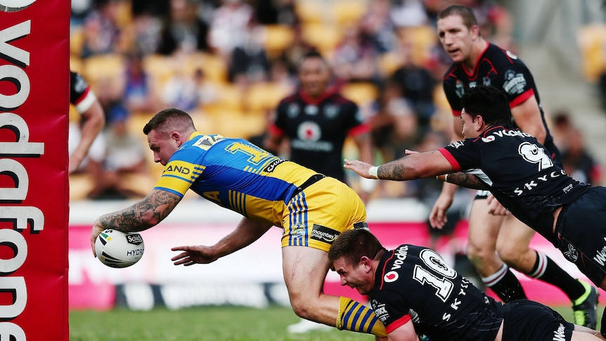 A Parramatta NRL player reaches out with the ball to score as he is tackled from behind by Warriors defenders.