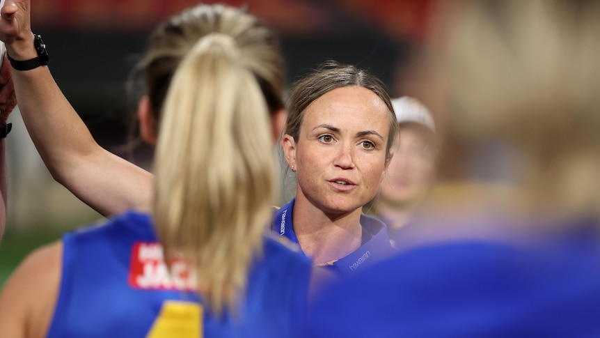 A West Coast AFLW coach looks out at her players (who have their backs to the camera) during a team talk in a game.