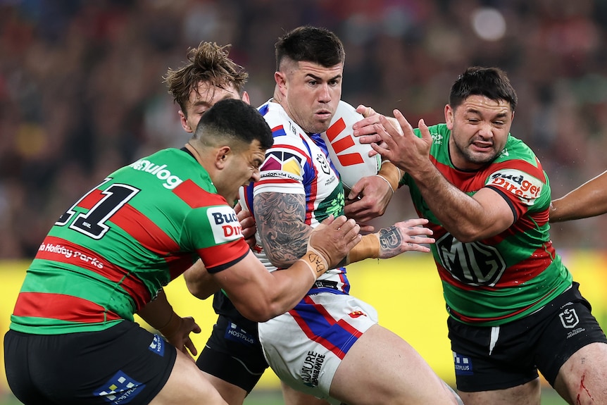 A group of rugby league players tackle for the ball, some in green and red and some in blue