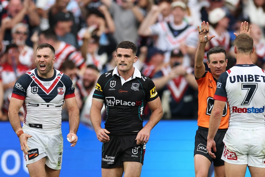 A rugby league player in black is pointed off the field by a referee in orange, opponents in white surround him