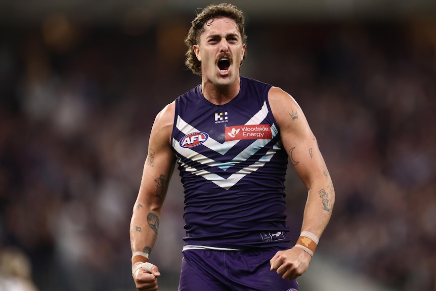 An AFL player in purple flexes his muscles in celebration of a goal