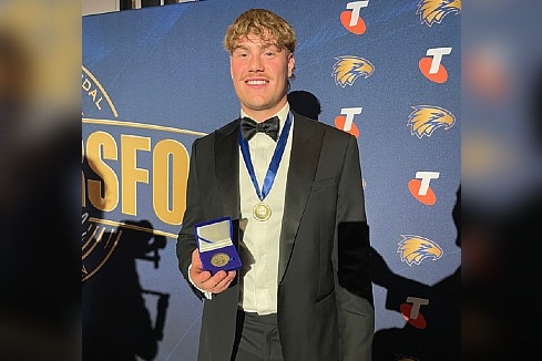 Harley wears a suit and smiles as he holds a medal at an awards ceremony.