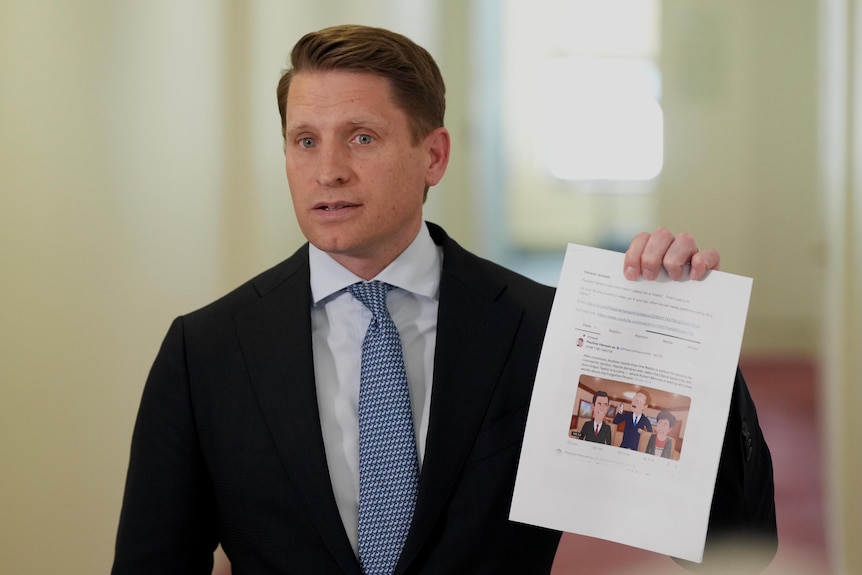 A white main with blue eyes and blue tie in suit looks annoyed and holds up a piece of paper with a screenshot of cartoon on it.