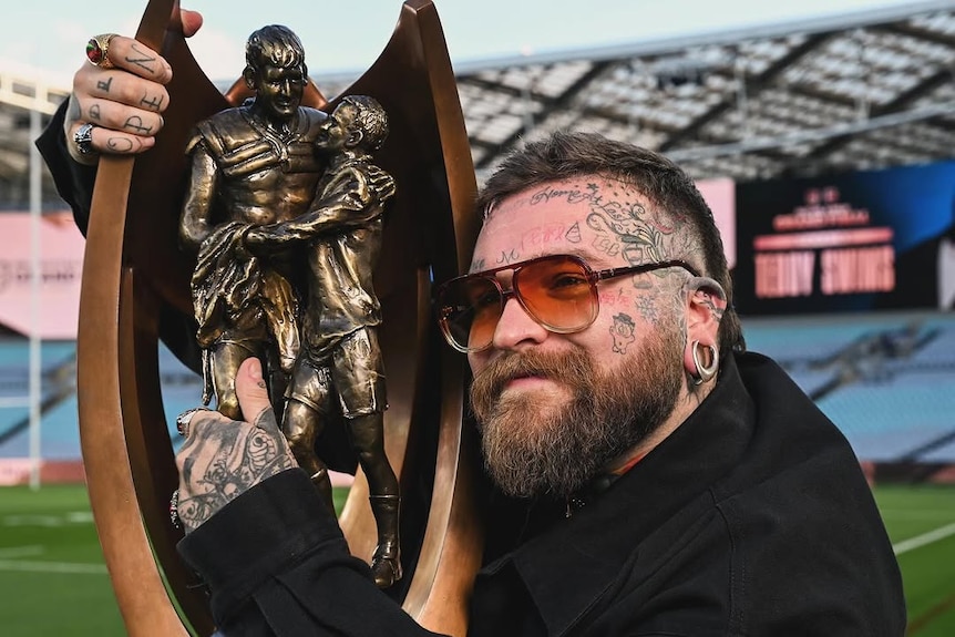 A dark-haired man with a face tattoo wears sunglasses as he holds a trophy in a football stadium.
