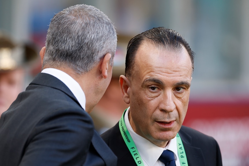 A grey-haired man speaks to a dark-haired man. Both wear suits.