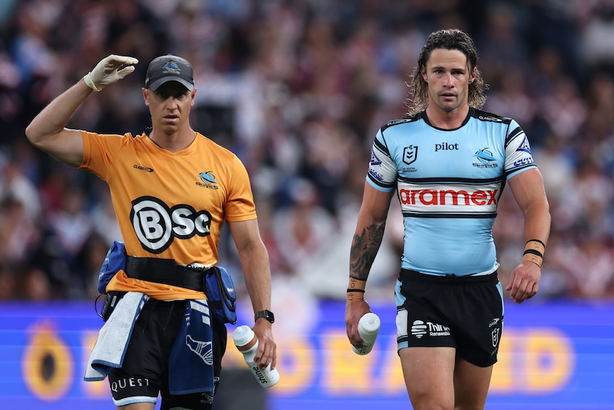 A rugby league player in blue limps from the field with a trainer next to him