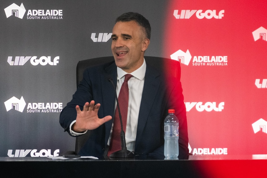 A man in a suit and red tie sitting at a table with LIV Golf signage
